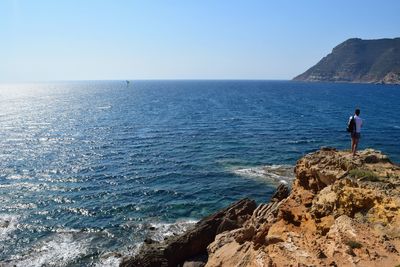 Rear view of man standing on cliff by sea against clear sky