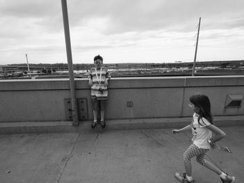 Sister running towards brother standing by retaining wall against sky