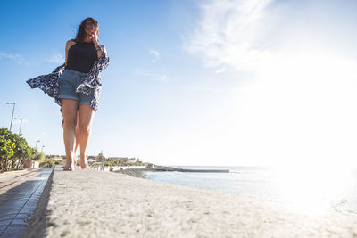 Low angle view of smiling young woman walking on retaining wall by sea