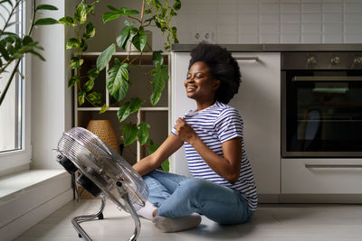 Happy satisfied african woman sits on floor in front of air cooler, cooling herself with ventilator