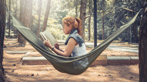Side view of woman reading book while sitting on hammock in forest