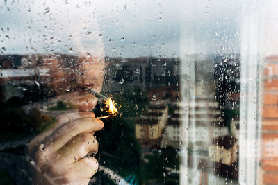 Unrecognizable male lighting cigarette with match while standing near wet window in rainy weather and spending day alone at home during quarantine because of coronavirus