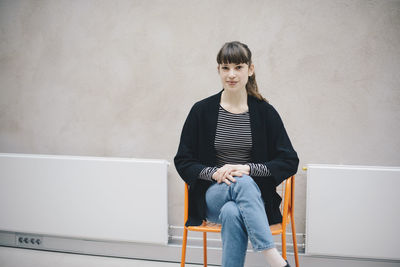Portrait of confident female computer programmer sitting on chair against beige wall in office