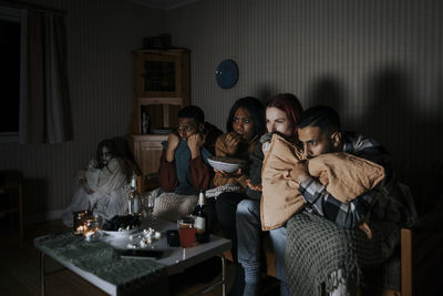 Frightened multiracial friends watching scary movie while ghost sitting in corner at cabin