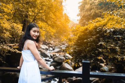 Portrait of smiling young woman standing by railing against trees