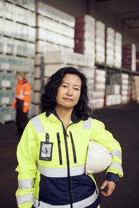 Portrait of confident female worker in reflective workwear holding hardhat standing in factory