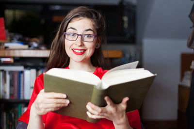 Smiling young woman wearing eyeglasses while reading book at home