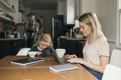 Woman using laptop while daughter having breakfast with family in background at home