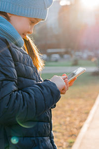 A girl in a jacket is holding a smartphone and texting a message in the rays of the evening sun