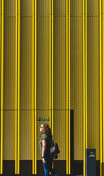 Side view of mature woman with backpack standing against yellow wall