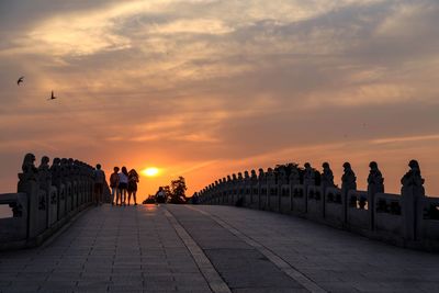 People on bridge against cloudy sky during sunset