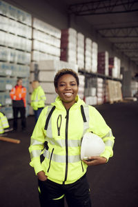 Portrait of smiling female worker in reflective clothing holding hardhat standing in factory