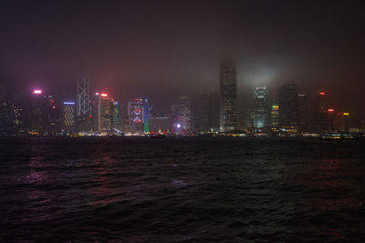 Illuminated buildings by sea against sky at night