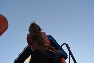 Low angle view of teenage girl with long hair standing on outdoor play equipment against clear sky