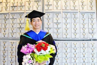 Portrait of young man wearing graduation gown standing with flowers against wall
