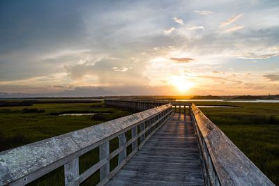 Boardwalk amidst field against sky during sunset