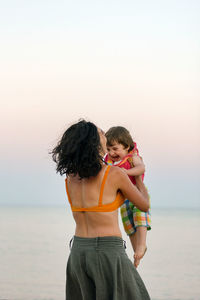 Rear view of mother and daughter standing on beach against clear sky