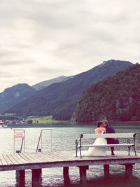 Rear view of bride and groom embracing while sitting on pier over lake against mountains