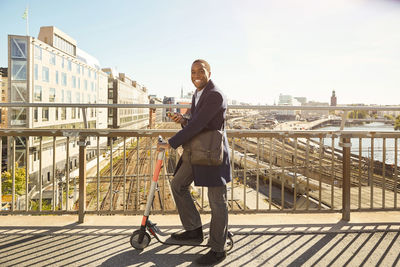 Side view portrait of smiling young man standing with electric push scooter on bridge in city