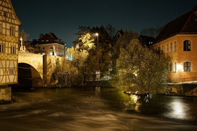 Illuminated bridge over river by buildings against sky at night