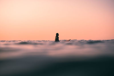 Rear view of woman sitting on a surfboard in the  ocean against an orange peach sky during sunset