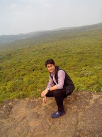 Full length side view portrait of young man crouching on mountain against sky