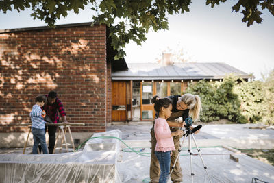 Mother talking to daughter by surveyor while son learning carpentry from father outside house