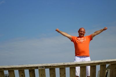 Portrait of man standing with arms outstretched by railing against sky