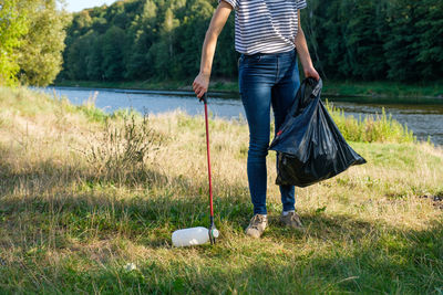 Low section of woman collecting garbage on land against river