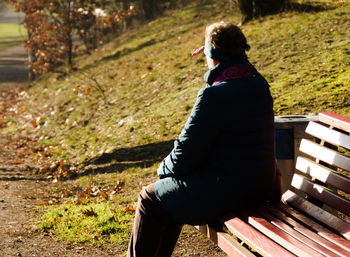 Rear view of woman shielding eyes while sitting on bench during sunny day at park