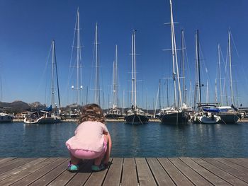 Rear view of baby girl crouching on pier at harbor with boats in background against clear sky
