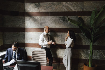 Business colleagues discussing while leaning on wall near businessman working at desk in office