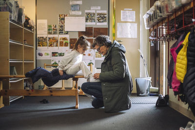 Side view of female teacher helping girl while tying shoe lace in cloakroom at kindergarten