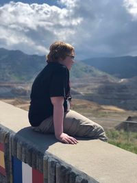 Side view of overweight boy looking at landscape while sitting on retaining wall against sky