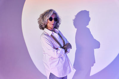 Elegant elderly female in white shirt and sunglasses crossing arms while standing in bright spotlight circle and looking at camera