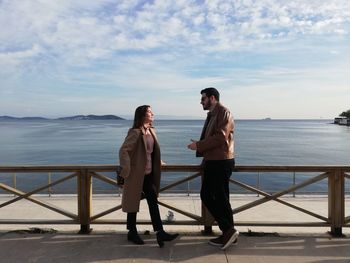 Full length of friends talking while standing by railing against sea