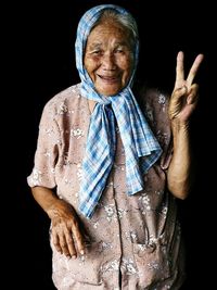 Portrait of smiling senior woman gesturing peace sign while standing against black background