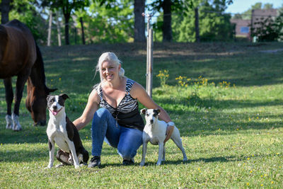 A young blonde pretty woman kneeling on lawn with her two dogs to her side looking to the camera.