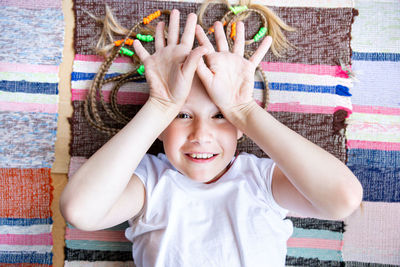 A cheerful girl with pigtails, lies on a woven striped rug, smiles, shows open palms 