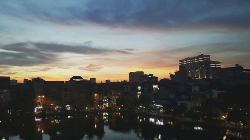 Illuminated buildings by river against sky at sunset