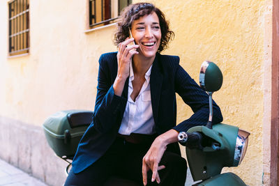 Cheerful female in formal outfit laughing brightly while sitting on motorcycle and talking on cellphone near stone building