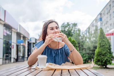 Young woman drinking coffee eating burger while sitting in street cafe