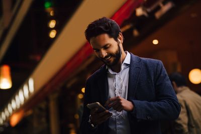 Low angle view of smiling man using smart phone with in-ear headphones while standing in city at night
