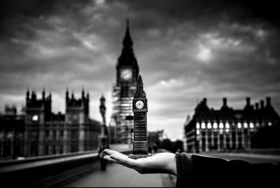 Cropped hand holding big ben model in city against cloudy sky