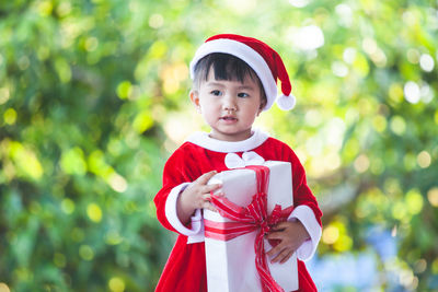 Cute baby girl wearing santa costume holding christmas present while standing against trees