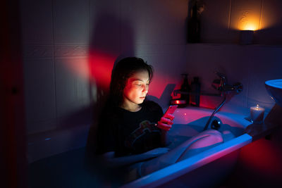 Young woman using smartphone while sitting in bathtub full of water, suffering from phone addiction