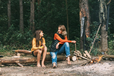 A mother with her teenage daughter is sitting on a tree near a campfire in the forest 