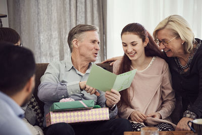 Grandfather reading greeting card to happy family while sitting on sofa at home