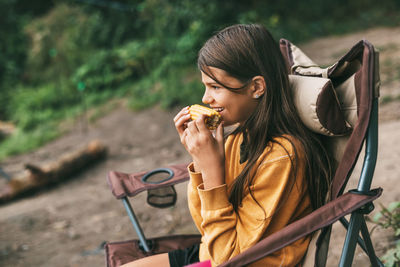 A teenage girl in a bright yellow sweater is sitting in a camping chair on the shore of the lake