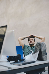 Thoughtful male computer programmer sitting with hands behind head and feet up at desk against beige wall in office
